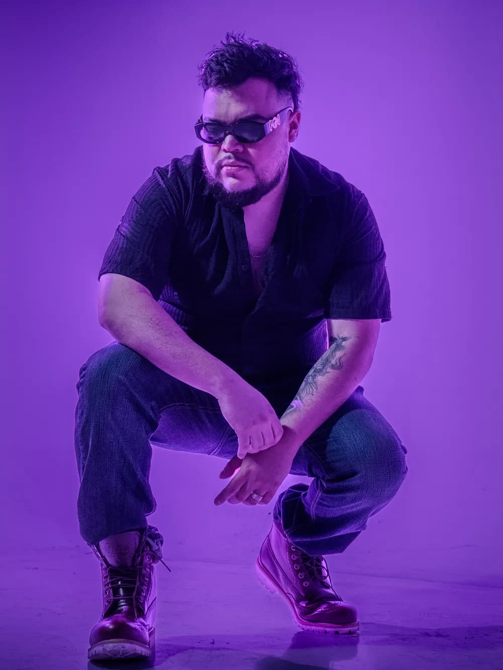 Papi AQ crouched in sunglasses against purple light.