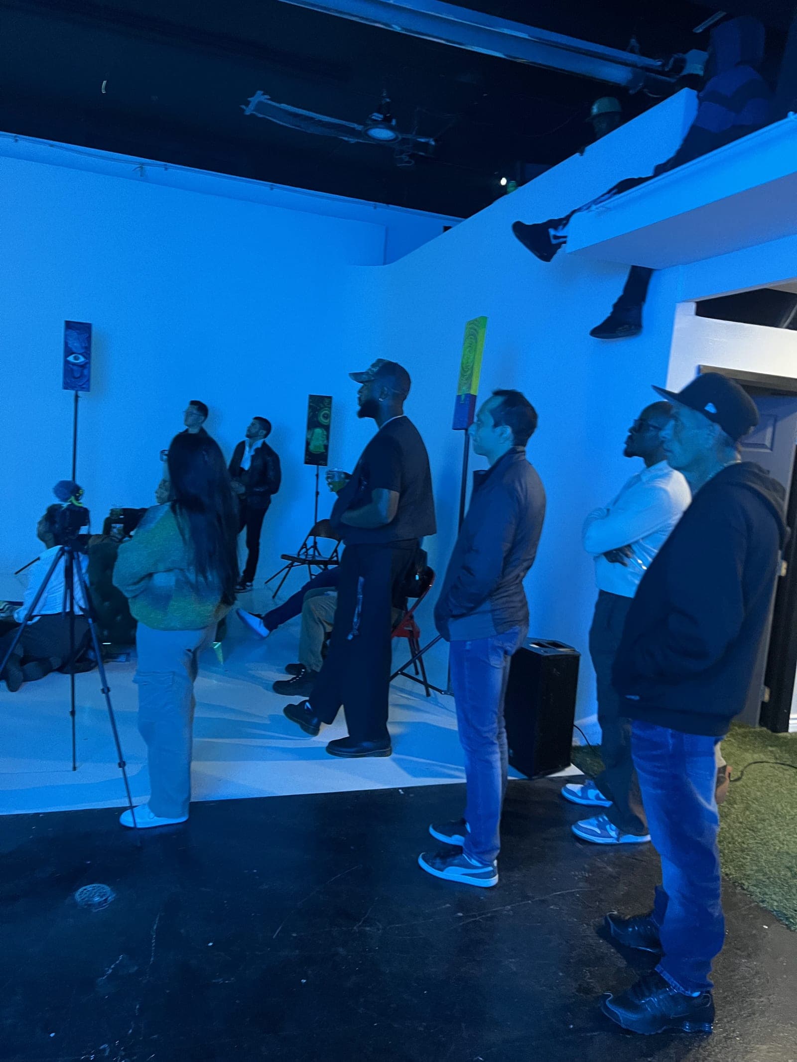 Guests stand in a room lit blue, all looking up at a screen out of frame. One guest sits high on a ledge above the doorway with their legs hanging over the edge.