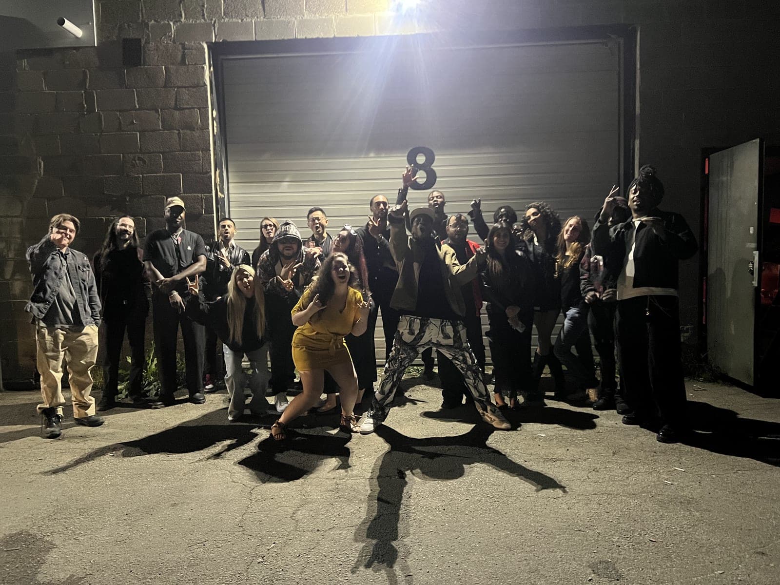 About thirty people pose together at night in front of a garage door marked with a large number 8, lit by one floodlight. Hands are up in signs and cheers, a guest in a yellow dress crouches low at the front and another in patterned trousers strikes a wide lunge.