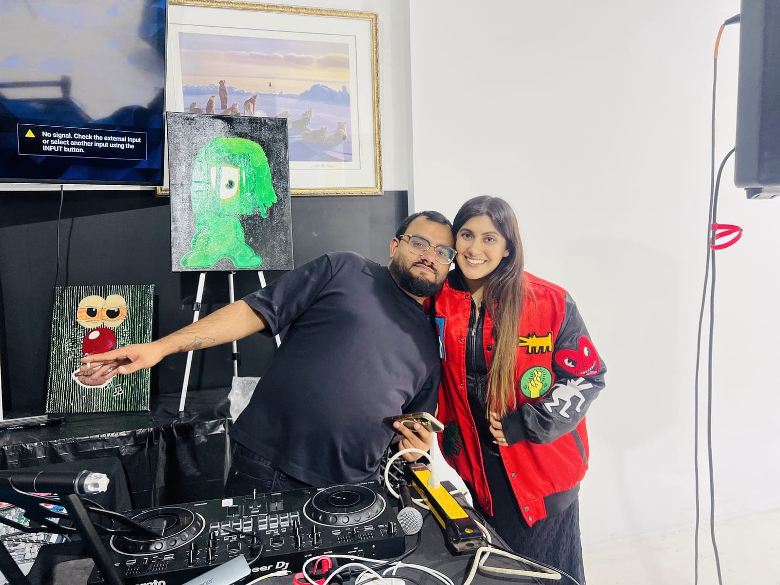 A DJ and a guest in a red jacket pose together at the decks, pointing at a painting of a green one-eyed character, with a small painting of a face with wide eyes below it.