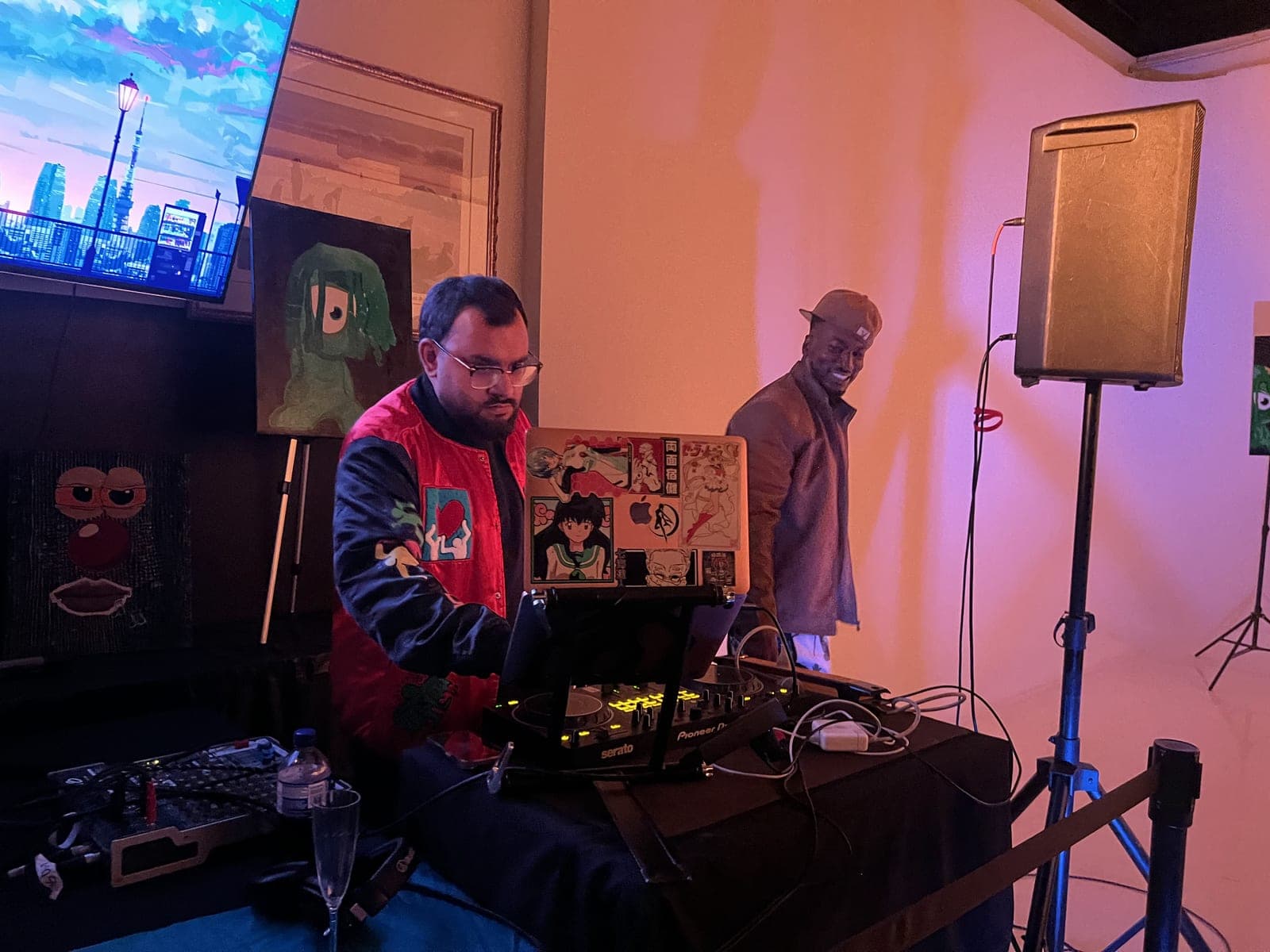 A DJ in a red and black jacket plays on a controller under warm orange light, beside a painting of a green one-eyed character, while another guest stands by the speaker.