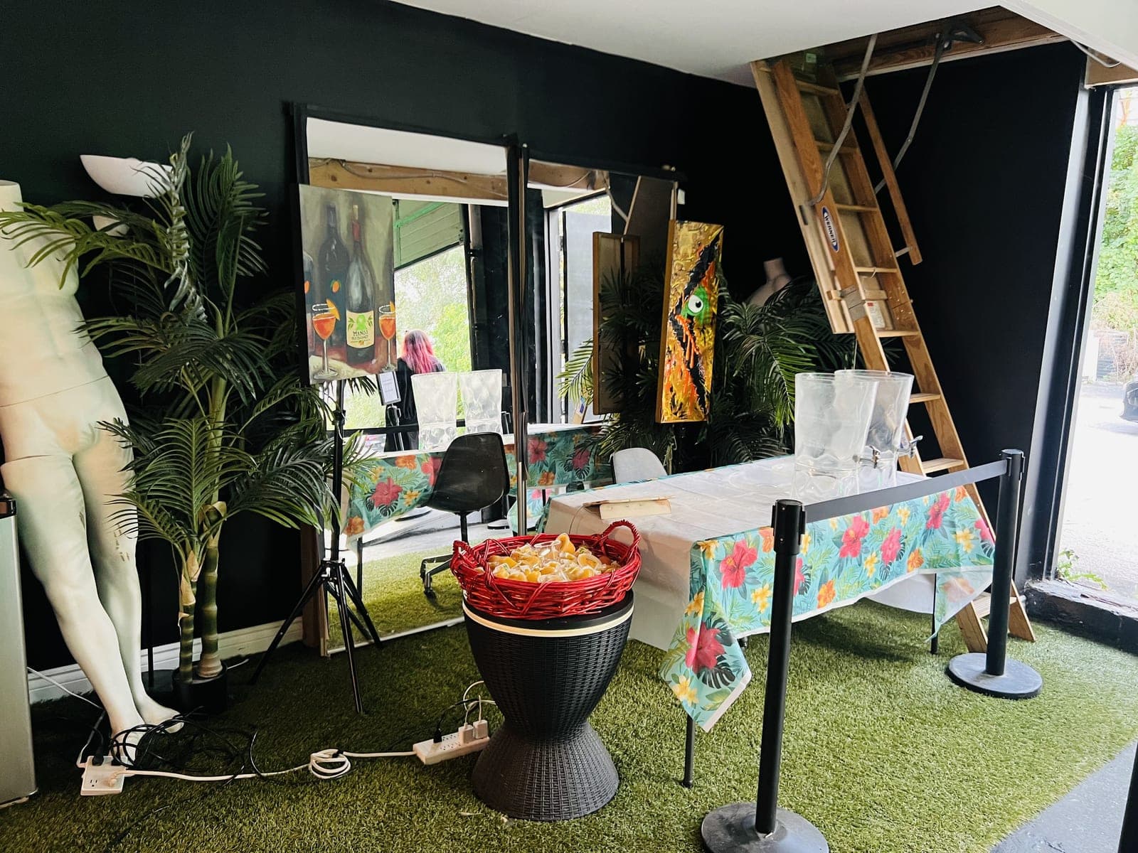 The entrance: artificial turf, potted palms, a white mannequin, a painting of two bottles on an easel, a red basket of mango cups and a table with a tropical print cloth beside a wooden loft ladder.