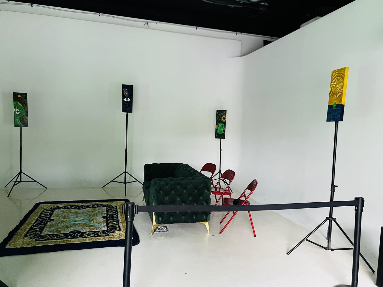 A white corner of the room with tall narrow paintings on stands, one showing a single eye, around a dark green velvet sofa, a patterned rug and red folding chairs.