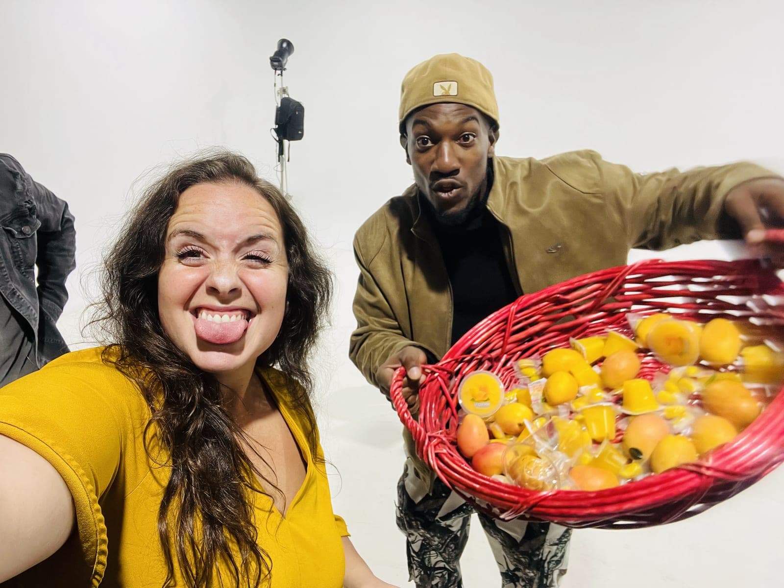 A guest in a yellow top sticks their tongue out in a selfie while another guest in a tan cap leans in behind, tipping a red basket of mangoes toward the camera.