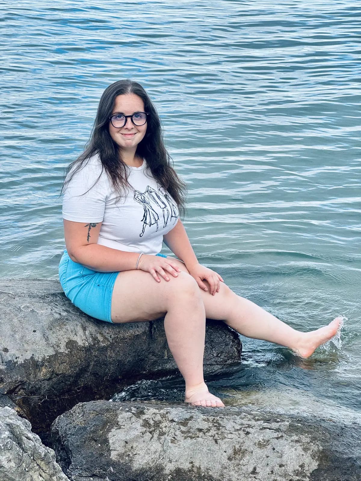 Amanda Andress sitting on a rock at the edge of the water, one foot in the lake, smiling at the camera.