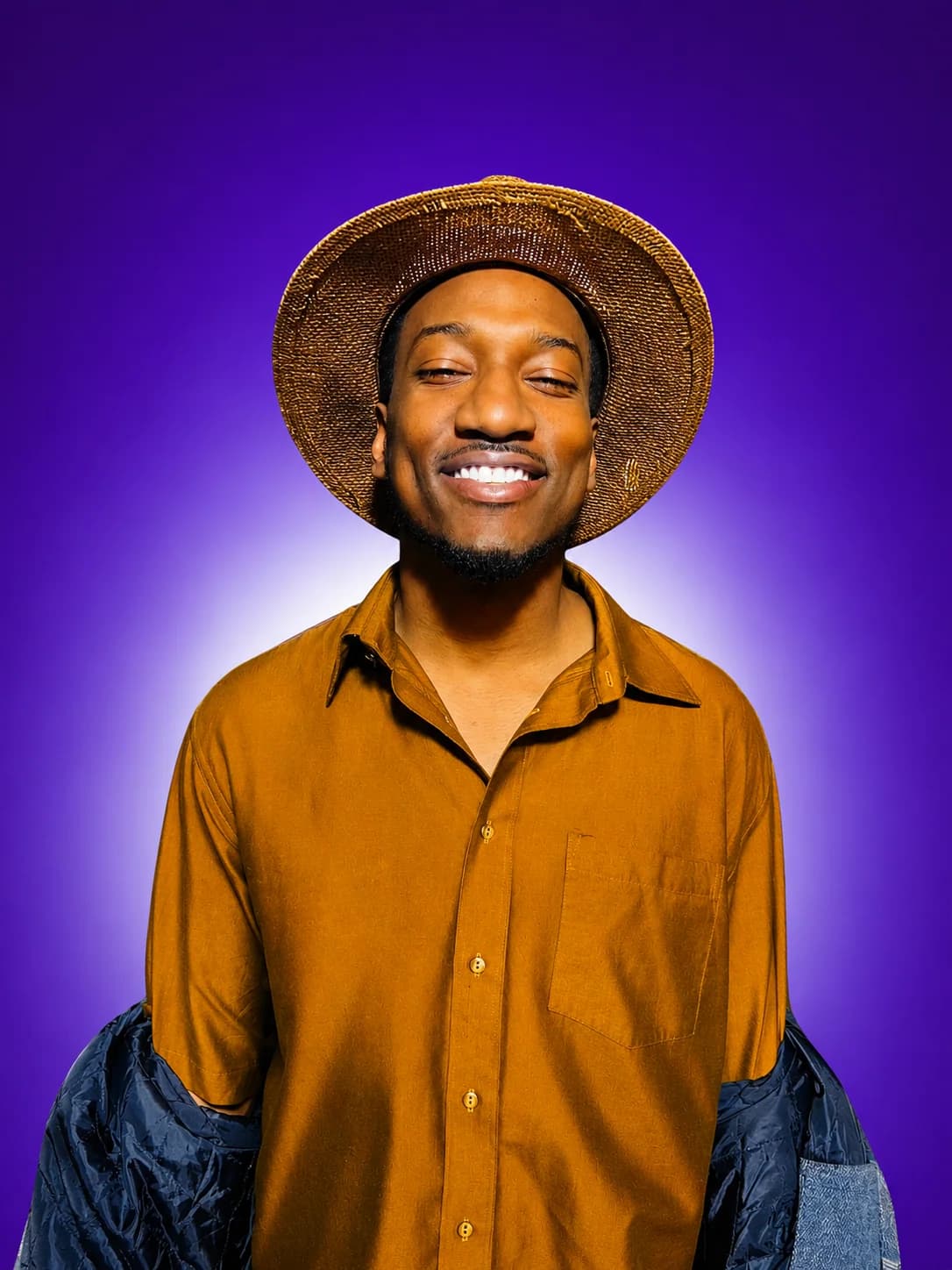 Colour press photograph of Cassius Crieghtney smiling in a straw hat and an orange shirt, lit against a purple background.