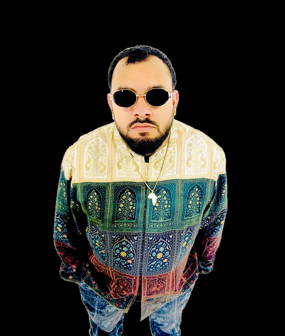 Sylus Rose in round gold sunglasses and a patterned jacket of cream, emerald, indigo and burgundy panels, looking up at the camera.