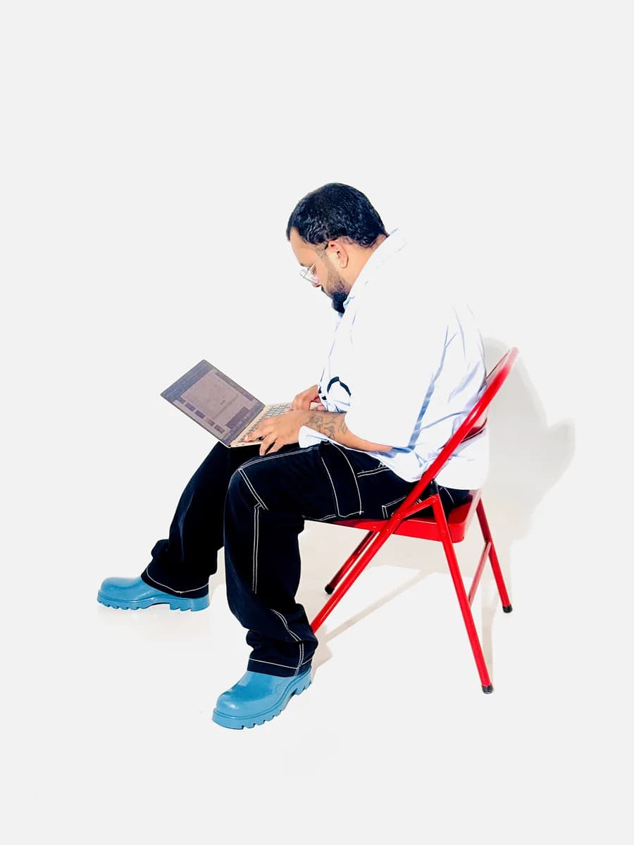 Sylus Rose on a red folding chair, working on a laptop, in a light blue shirt and teal boots.