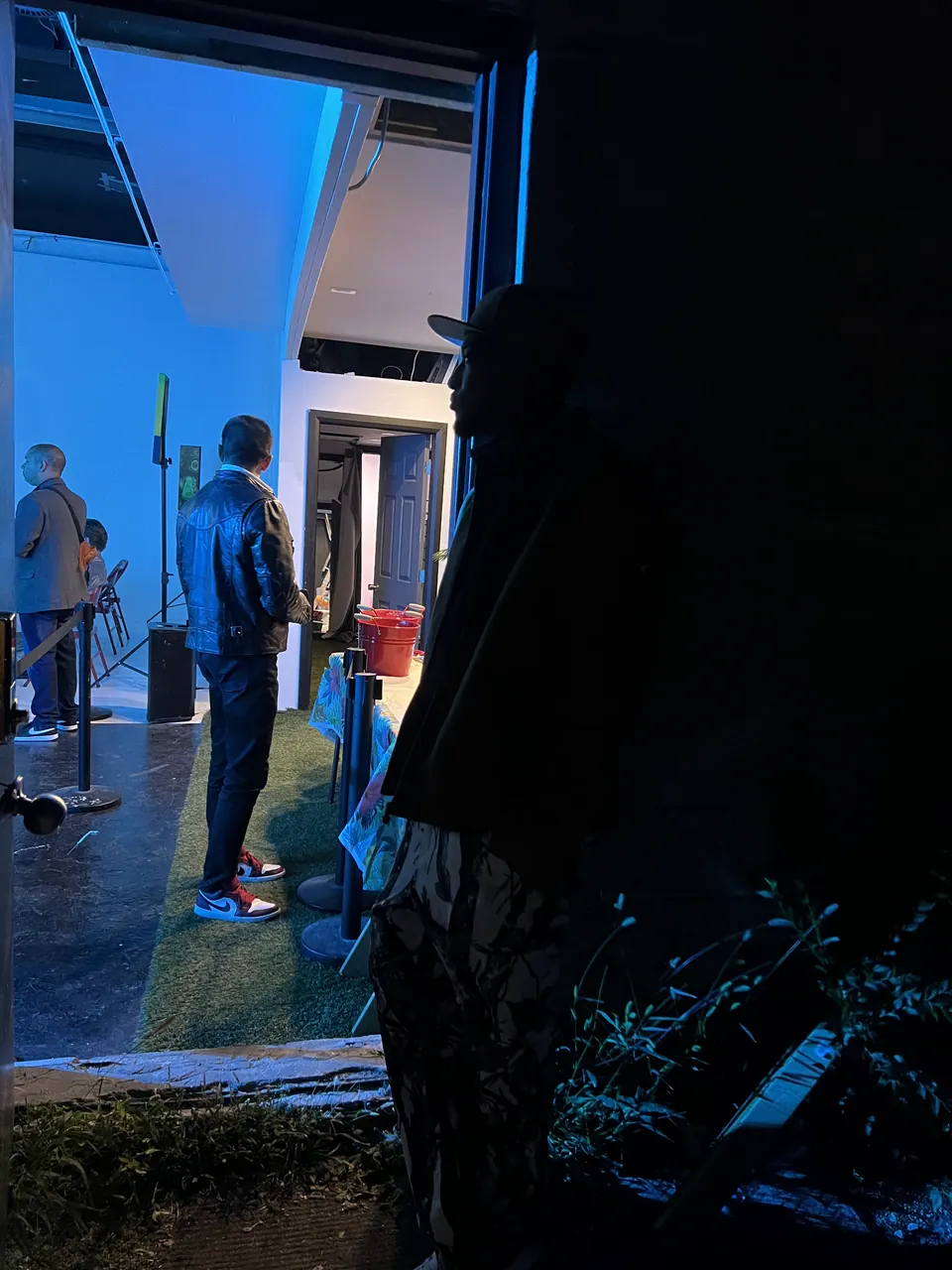 Looking through a doorway into a room lit blue where guests stand near a speaker, with a dark silhouette in the foreground.
