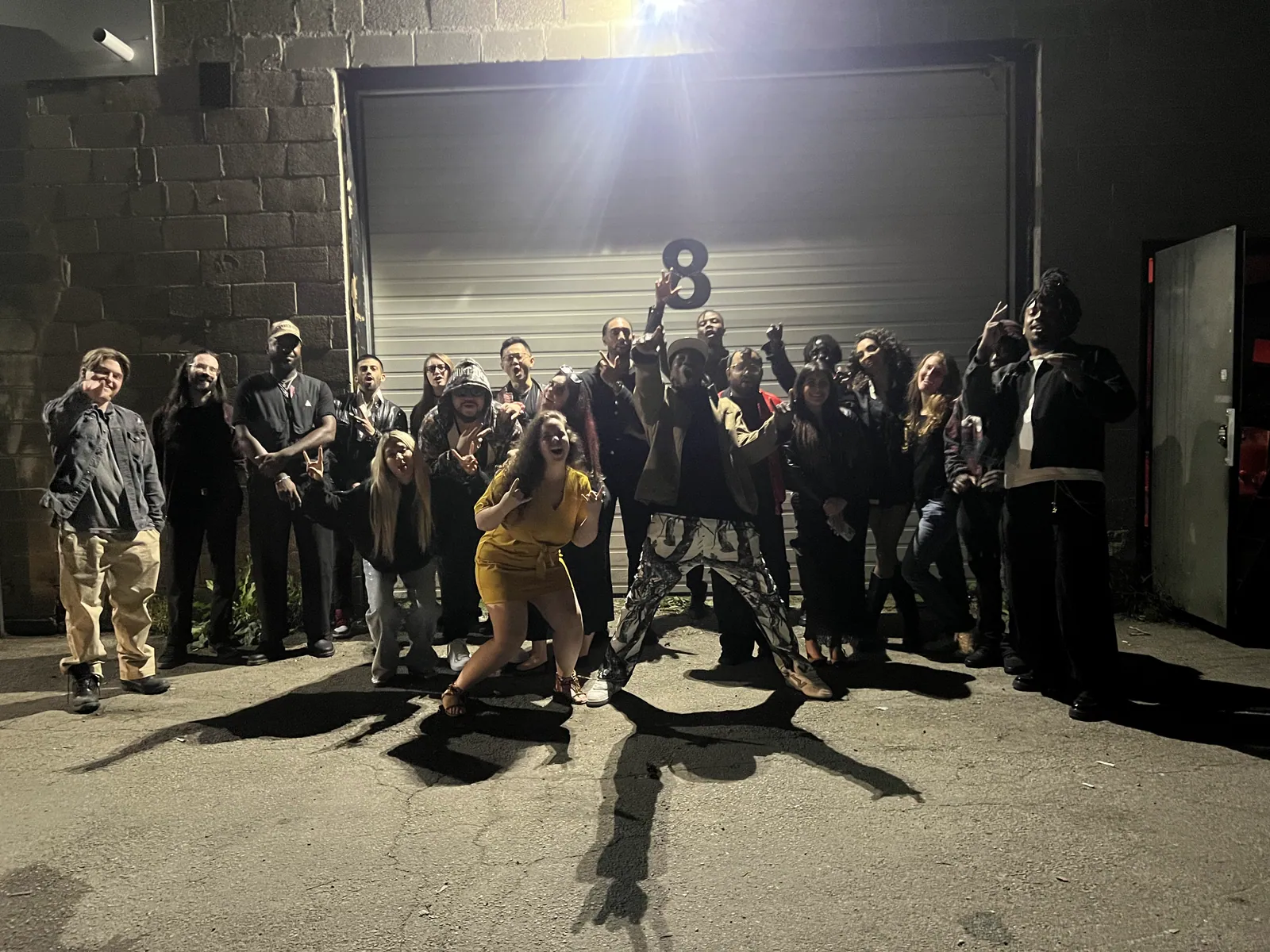 About thirty people pose together at night in front of a garage door marked with a large number 8, lit by one floodlight. Hands are up in signs and cheers, a guest in a yellow dress crouches low at the front and another in patterned trousers strikes a wide lunge.