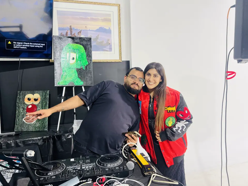 A DJ and a guest in a red jacket pose together at the decks, pointing at a painting of a green one-eyed character, with a small painting of a face with wide eyes below it.