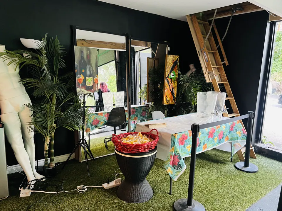 The entrance: artificial turf, potted palms, a white mannequin, a painting of two bottles on an easel, a red basket of mango cups and a table with a tropical print cloth beside a wooden loft ladder.