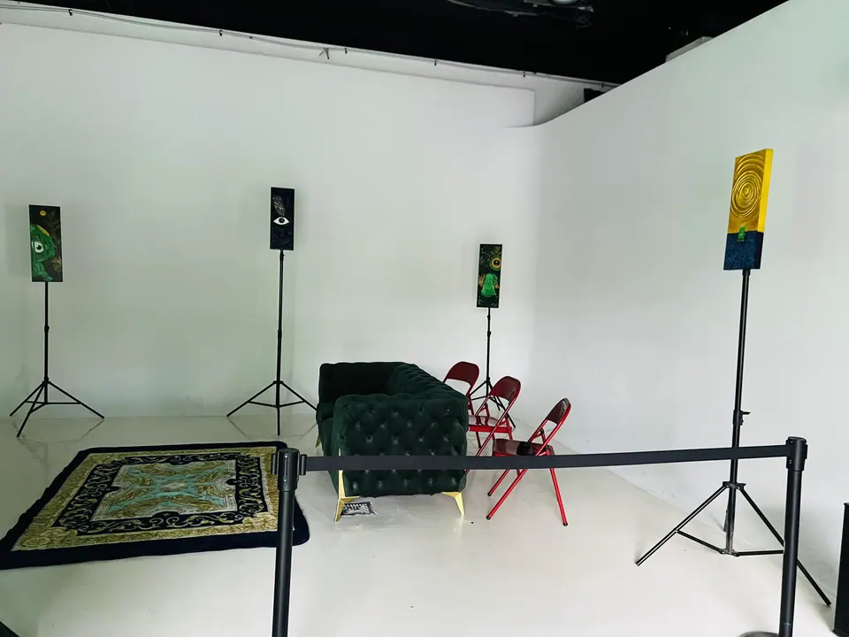 A white corner of the room with tall narrow paintings on stands, one showing a single eye, around a dark green velvet sofa, a patterned rug and red folding chairs.