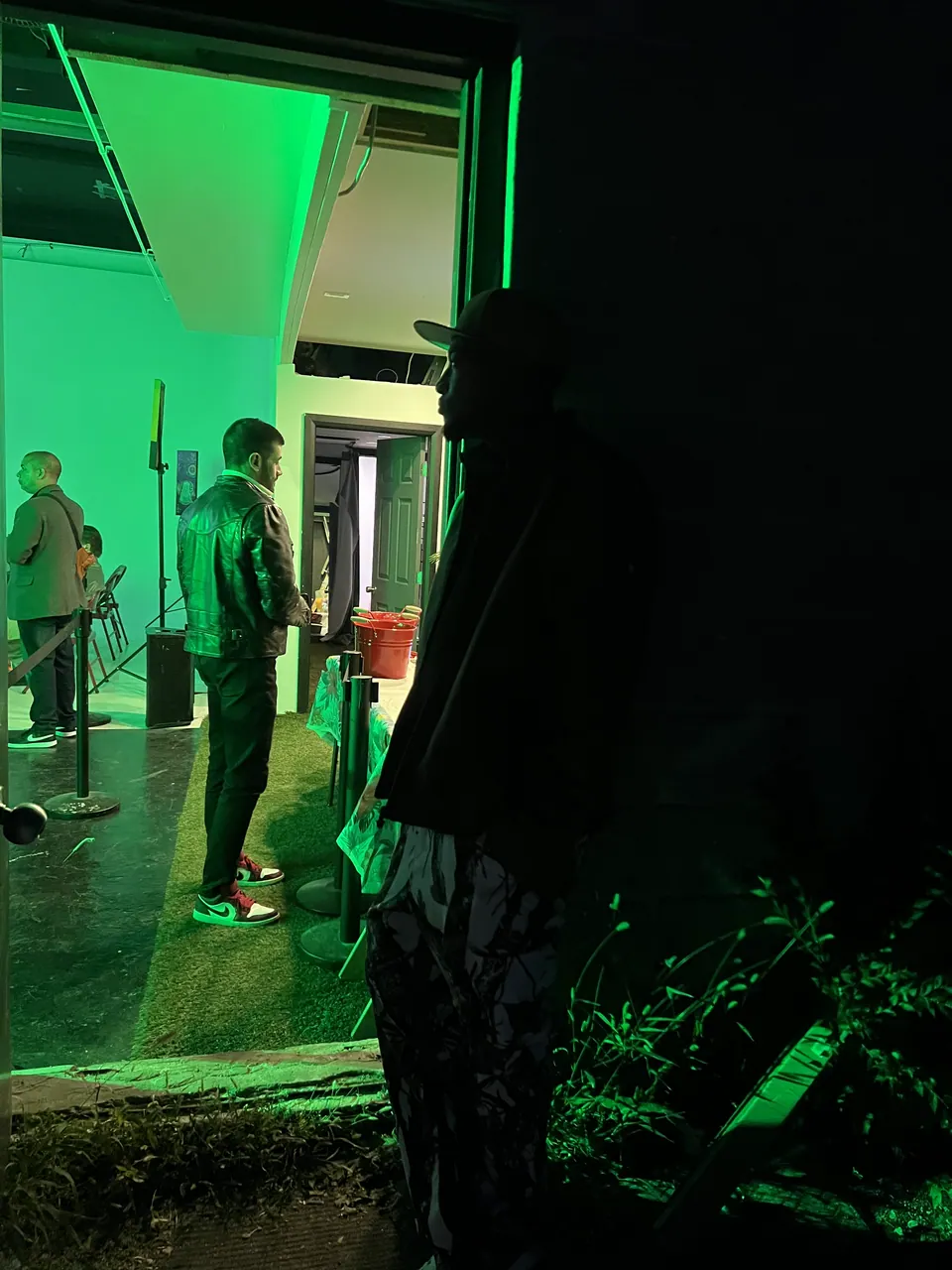 At night, a figure in silhouette stands in a doorway looking into a room washed in green light where guests stand and talk.