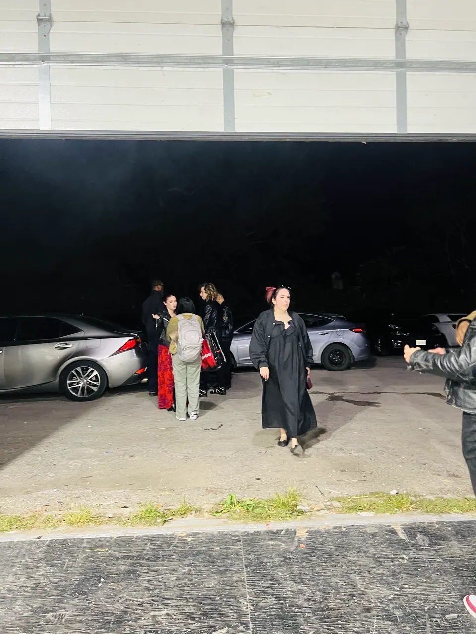 Seen from inside the open garage door at dusk, guests gather in the parking lot between parked cars.