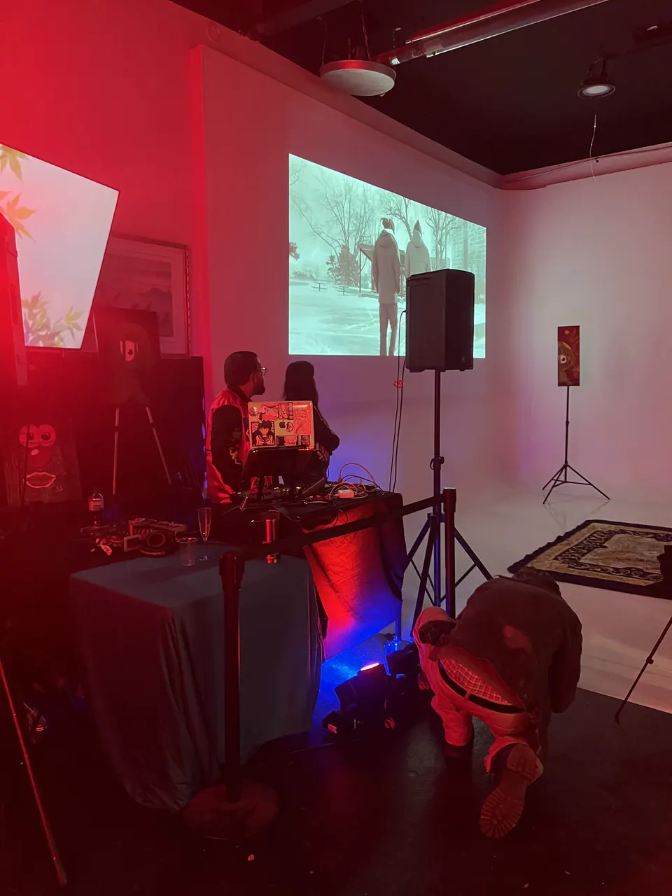 Under red light, a film scene of two figures in a snowy park is projected onto a white wall. Two people sit at the DJ table in front of it, and a guest crouches by the stage lights.