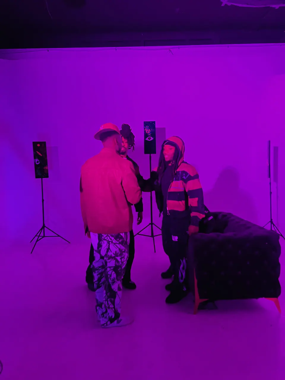 Three guests talk in a room lit purple, among speakers and paintings on stands.
