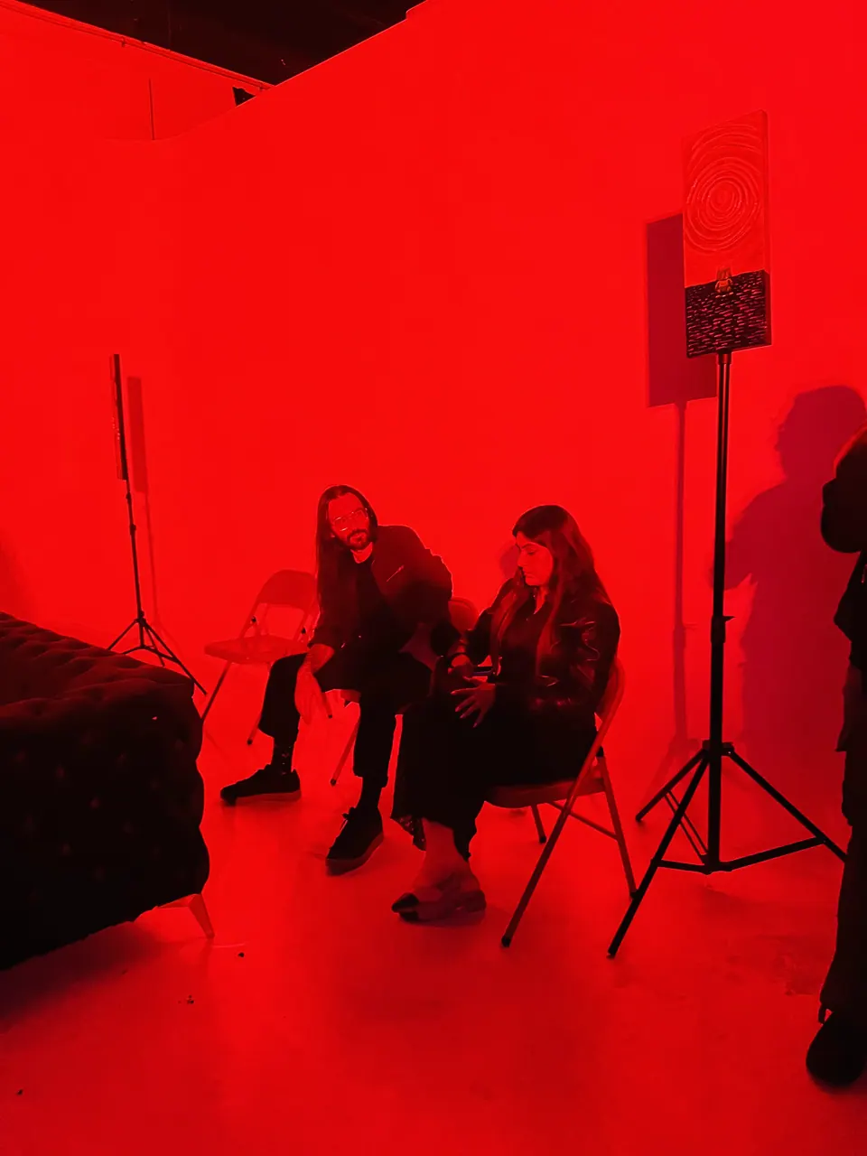 Two guests sit on folding chairs in a room flooded with red light, beside a painting on a stand.