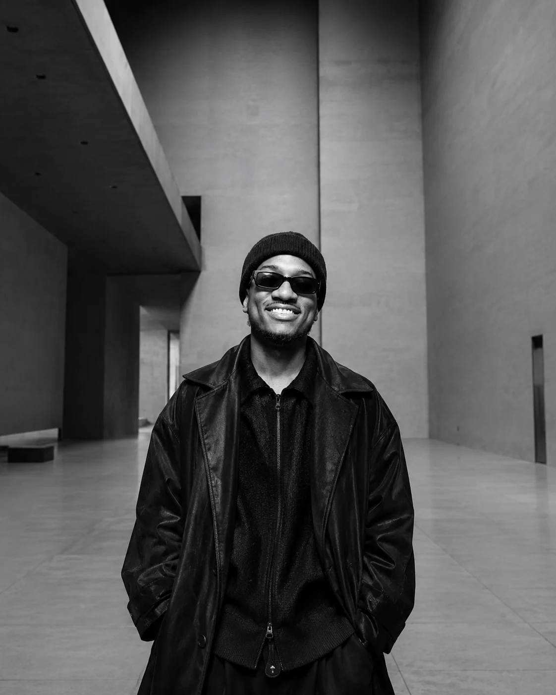 Black-and-white photograph of Cassius Crieghtney standing in a concrete gallery hall, in a leather jacket and sunglasses.